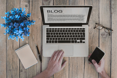 An overhead shot shows a laptop displaying the word "BLOG", a notebook, pen, phone, glasses, and a blue plant on a wooden surface.